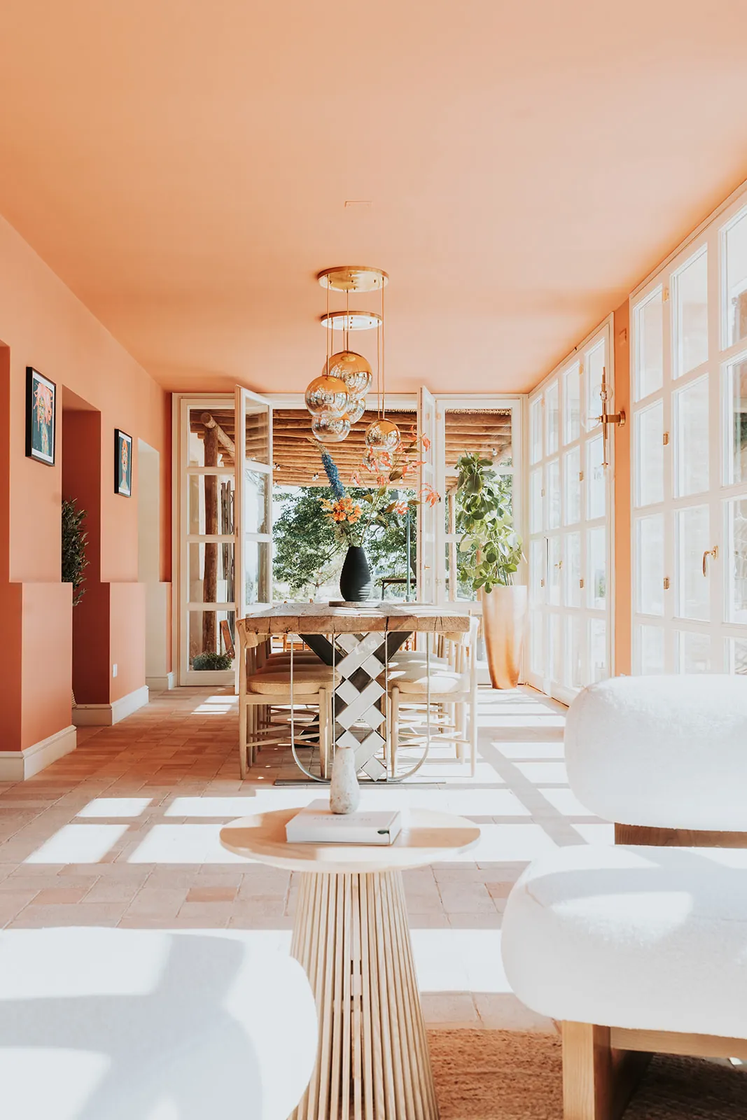 Glass house dining room with peach walls, a long wooden table and brass pendant lights