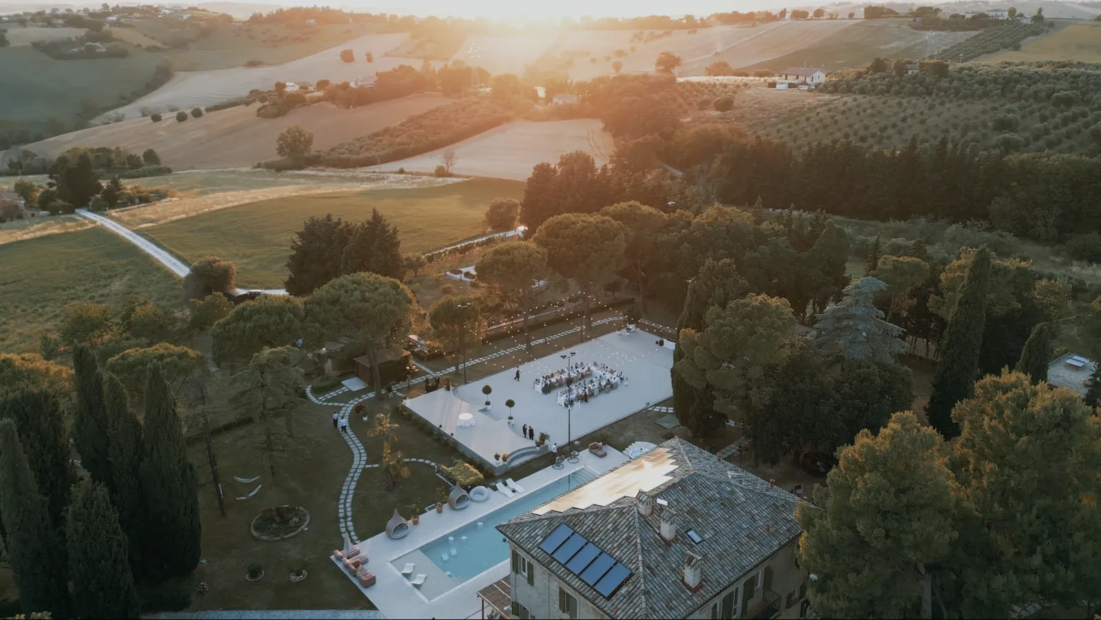 Drohnenaufnahme von Maniero Montecassiano bei Sonnenuntergang, mit gedeckter Tafel auf dem Rasenplatz oberhalb von Pool und Villa