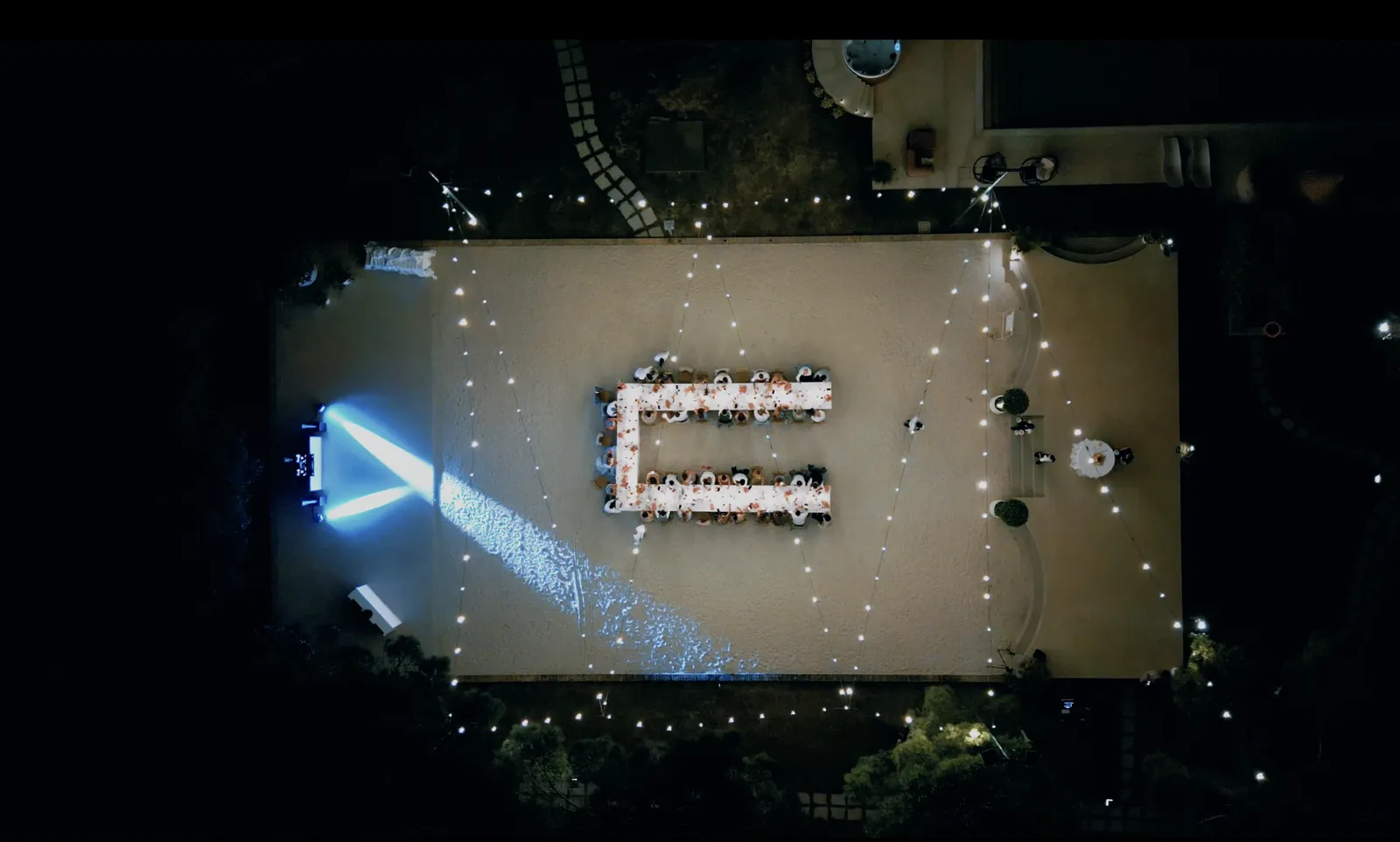 Nächtliche Drohnenaufnahme der Hochzeitstafel unter Lichterketten, mit den Lichtstrahlen des DJs